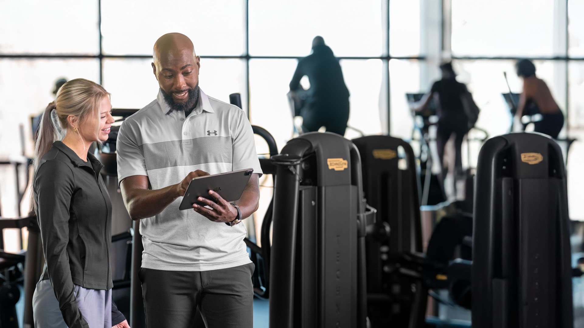 A fitness coach and a woman in athletic wear smile while looking at a tablet together in a gym.